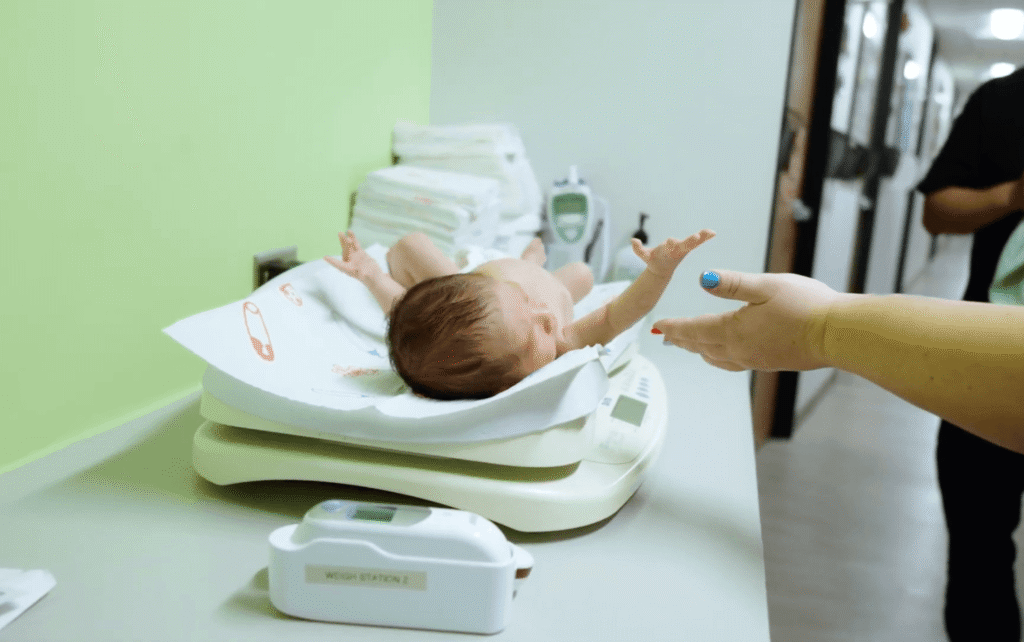 A newborn on an infant scale reaching toward an adult’s hand