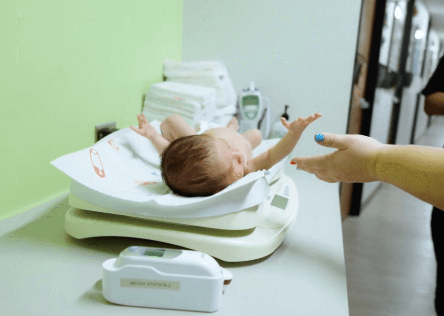 A newborn on an infant scale reaching toward an adult’s hand